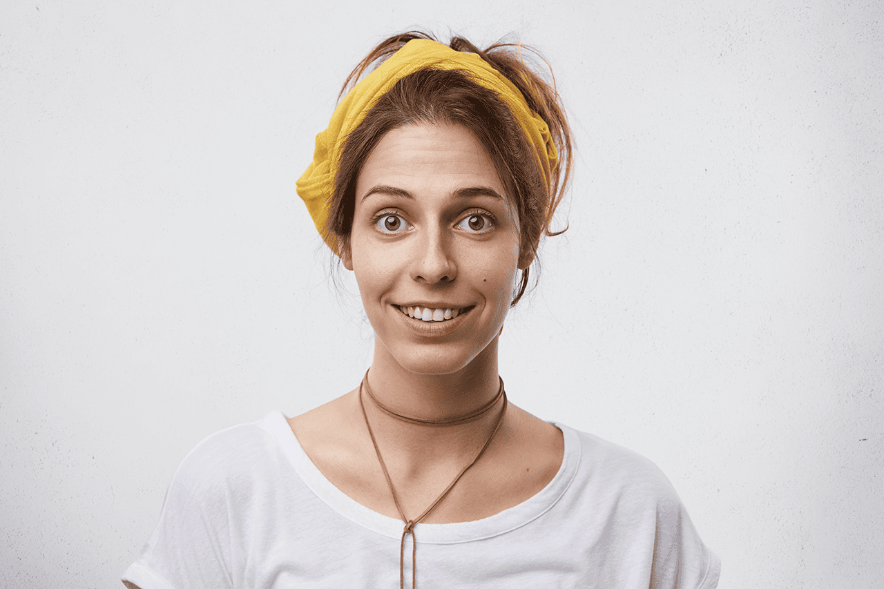 Jonge vrouw met een gele haarband die verwonderd en lachend in de camera kijkt
